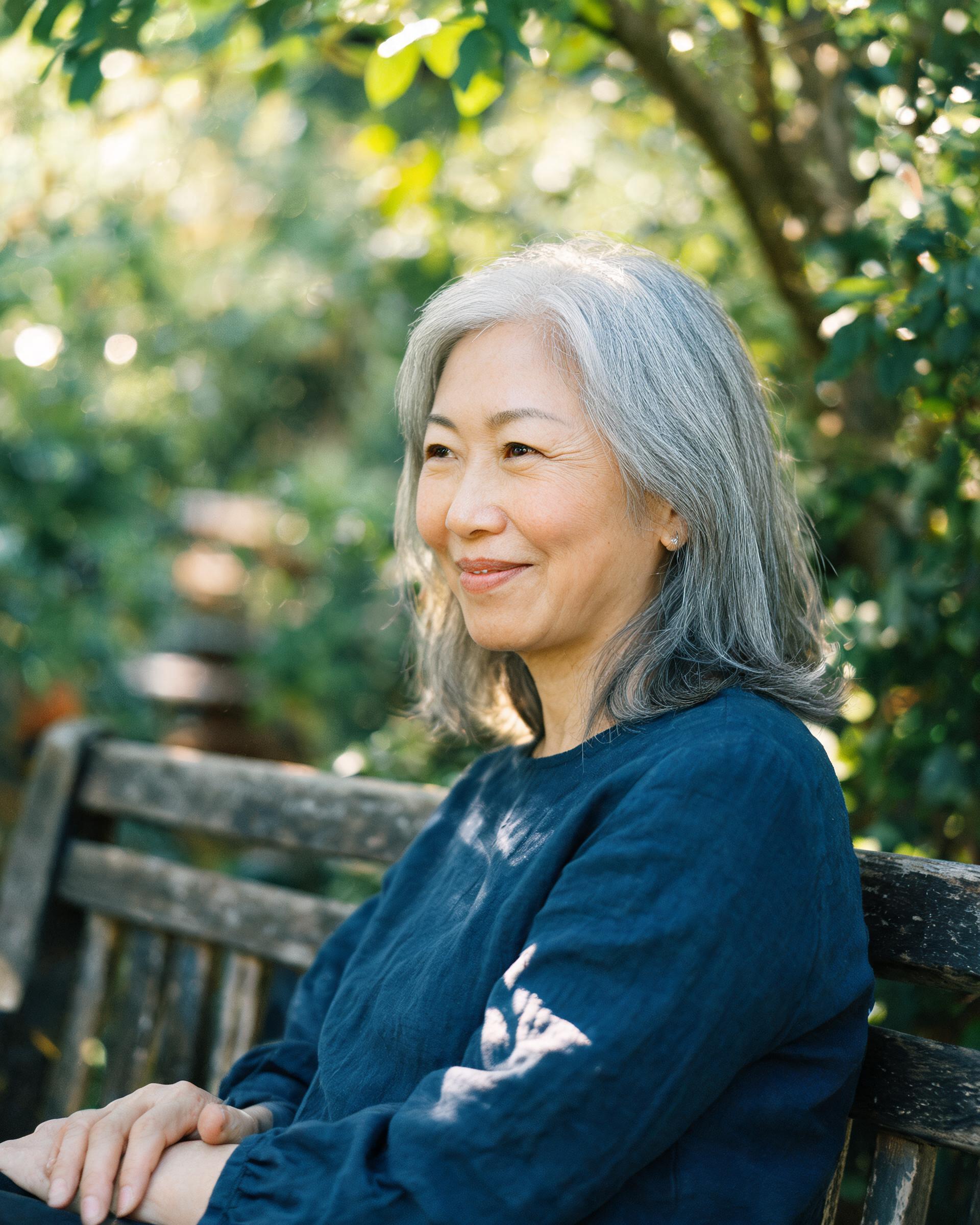 Mei W., a 56-year-old woman with silver hair, sitting on a garden bench